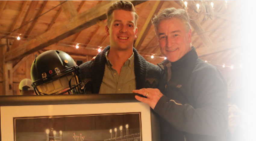 Two individuals posed in front of memorabilia including such items as portrait and football helmet.