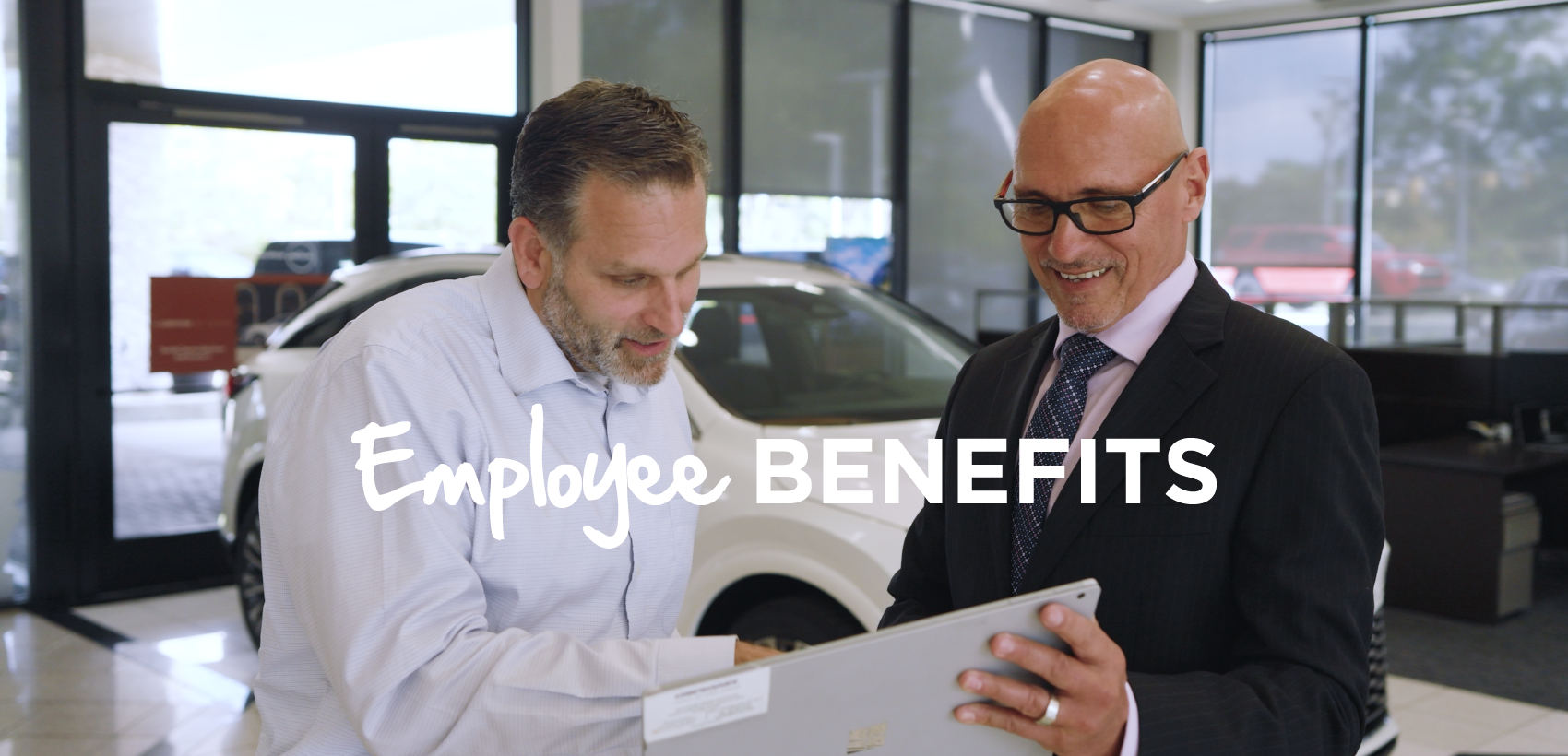 Text reads, “Employee Benefits” Above the text, two people in a showroom review information on a tablet beside a vehicle.