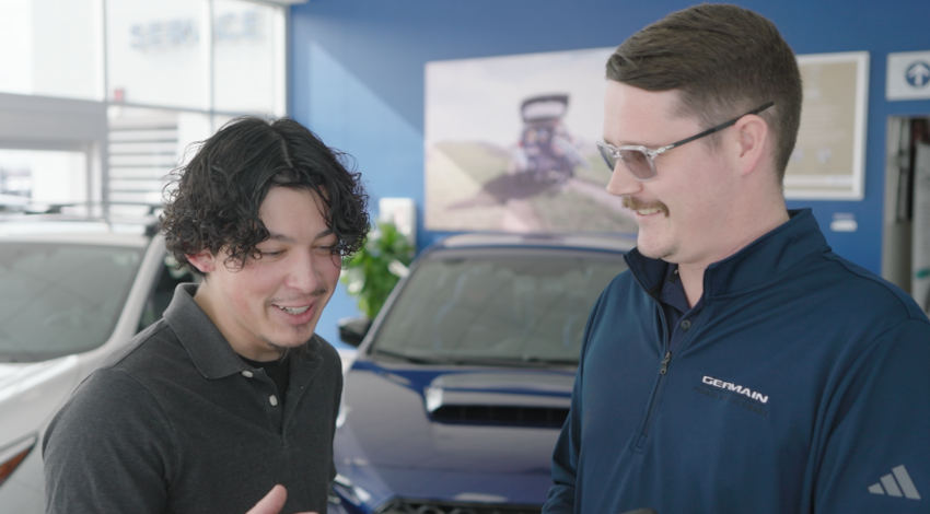 Two people stand beside vehicles in a showroom, looking at a smartphone together and smiling during a conversation.