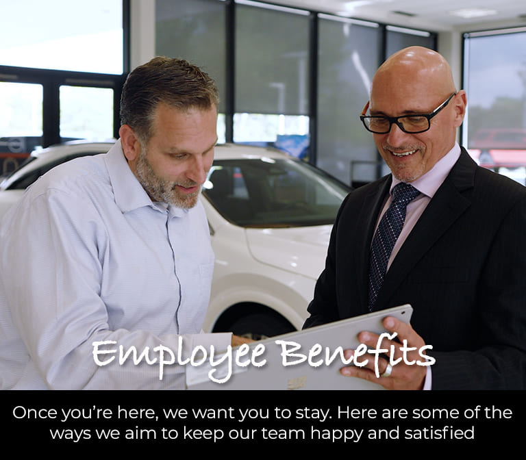 Two Germain employees reviewing information on a tablet in a dealership showroom. Text reads: "Employee Benefits – Once you're here, we want you to stay. Here are some of the ways we aim to keep our team happy and satisfied."
